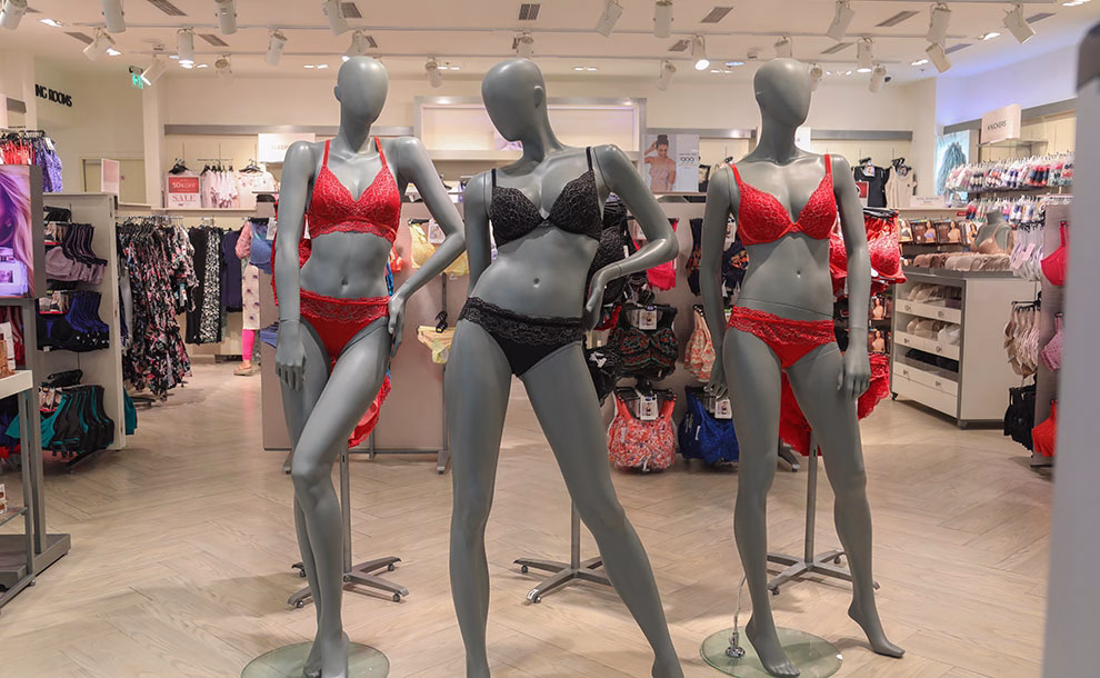 Photograph of three gray mannequins in a store, each wearing a bra and underwear set. The mannequins on the left and right wear red sets, while the center mannequin wears a black set. Racks of clothing are visible in the background.